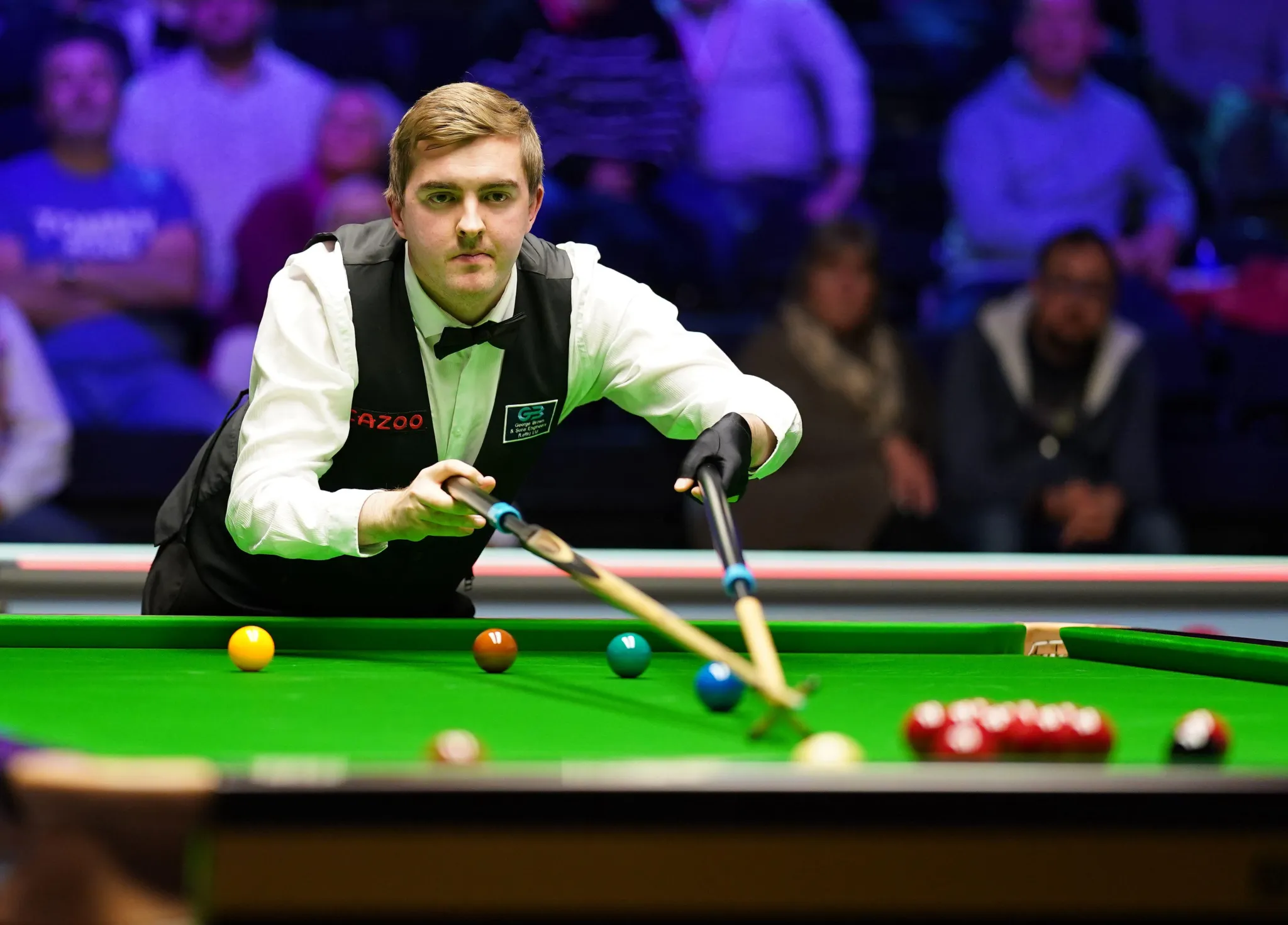 Ross Muir stretched over the table during a match, playing a shot with the audience out of focus beyond him.